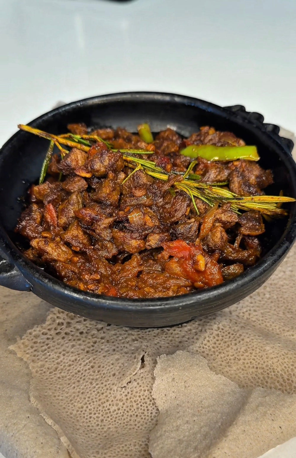 Awaze tibs with rosemary and green pepper in a black clay pot on injera