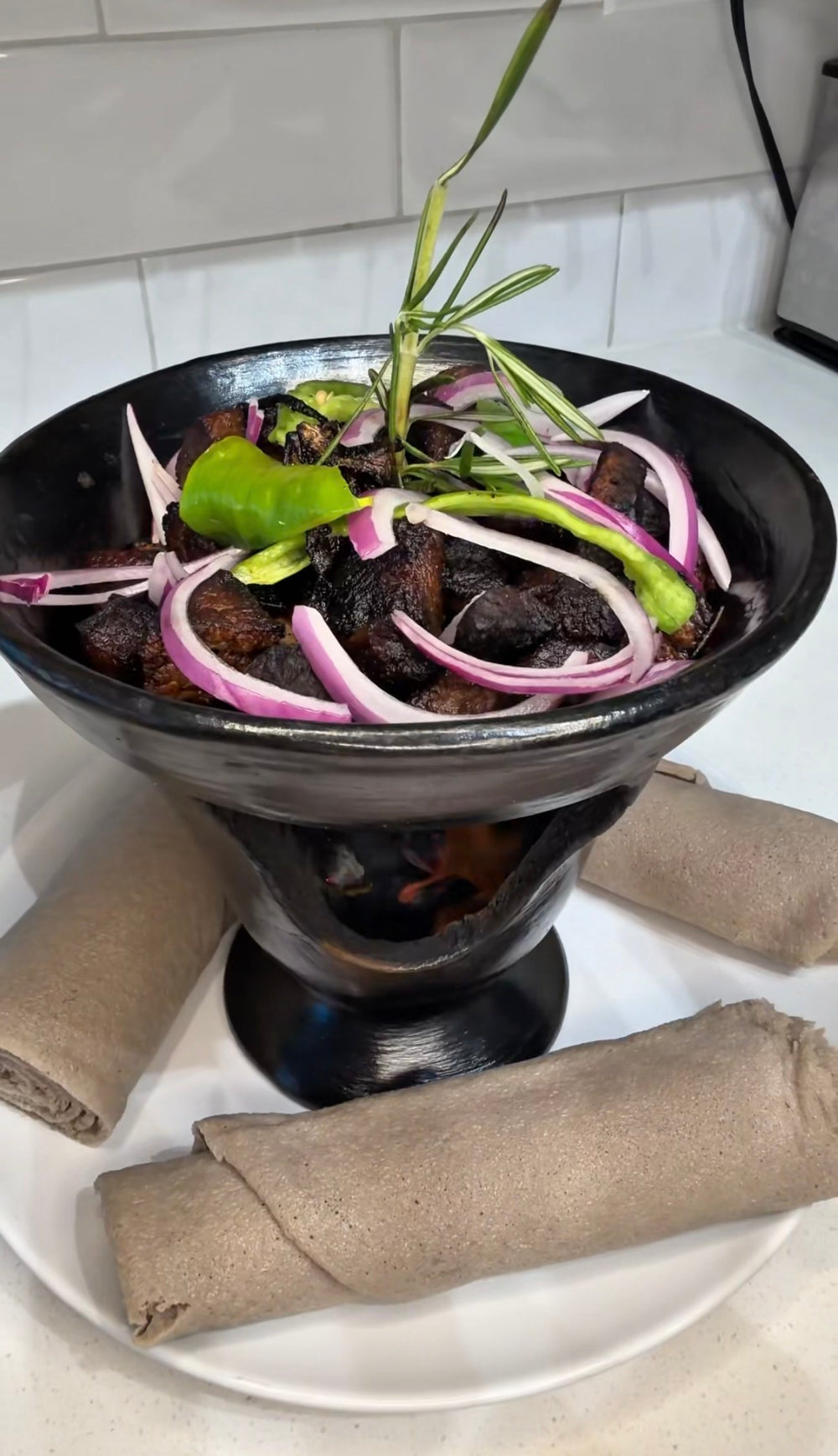 Shekla tibs in a clay shekla over coals, topped with sliced red onion, green pepper and rosemary, with rolled injera